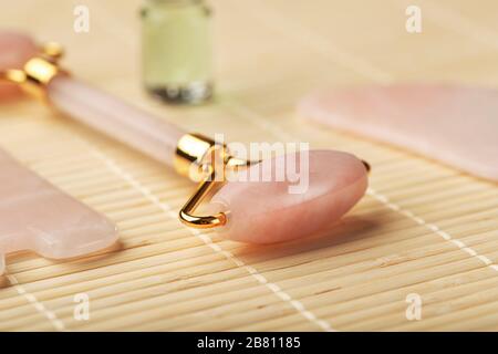 Eine Reihe von Werkzeugen für die Gesichtsmassage Gua Sha aus natürlichem Rosenquarz. Walze, Jadestein und Öl in einem Glasbecher, auf Strohgrund für Stockfoto