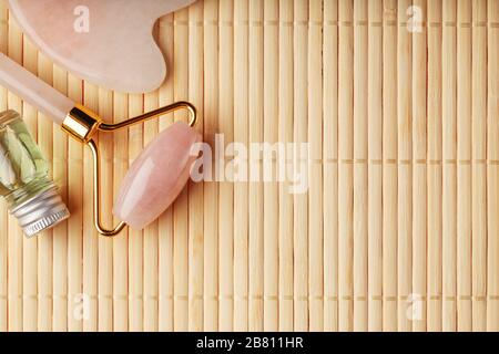 Eine Reihe von Werkzeugen für die Gesichtsmassage Gua Sha aus natürlichem Rosenquarz. Walze, Jadestein und Öl in einem Glasbecher, auf Strohgrund für Stockfoto