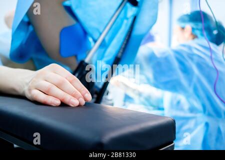 Hand eines Patienten, der auf dem Operationstisch liegt. Der Patient schläft unter Vollnarkose. Im Hintergrund sind Chirurgen in chirurgischer Kleidung a Stockfoto