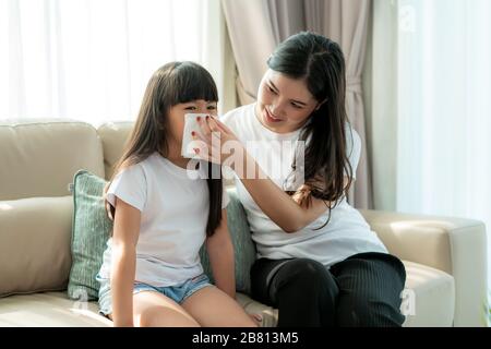 Porträt des geschnittenen asiatischen Mädchens, das mit ihrer Mutter in die Serviette bläst, hält es vorsichtig in der Nähe ihrer Nase. Bilder von Menschen, die sich krank und suff fühlen Stockfoto