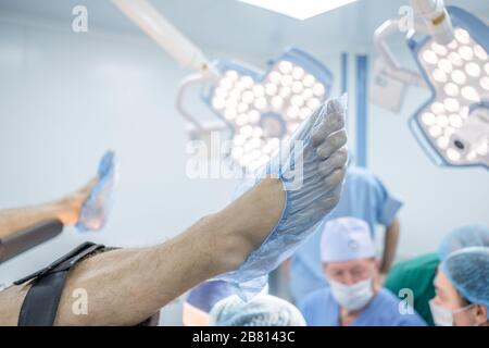 Die Beine des Patienten in medizinischen Schuhdeckeln liegen auf dem Operationstisch. Im Hintergrund sind Chirurgen in chirurgischen Kleidung und helle Operating l Stockfoto