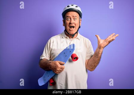 Mann im mittleren Alter, der einen Sicherheitshelm trägt, der das Skateboard über purpurem Hintergrund hält, sehr glücklich und aufgeregt, der Siegerausdruck feiert den Sieg Stockfoto