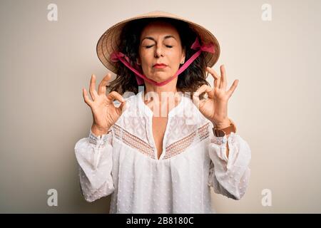 Frau im mittleren Alter mit asiatischem, traditionellen, konischen Hut über weißem Hintergrund entspannen Sie sich und lächeln Sie mit geschlossenen Augen, die mit Meditationsgeste tun Stockfoto