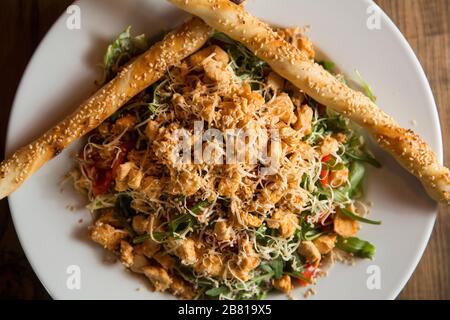 Hühnersalat mit Rakete, Kirschtomaten, Räucherkäse und knackigen Sesamstäbchen in der Platte auf dem Holztisch Stockfoto