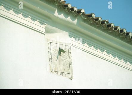Sonnenuhr mit römischen Ziffern, hoch oben an der weißen Steinwand eines Gebäudes in Parga, Preveza, Epirus, Griechenland angebracht. Das darüber liegende Ziegeldach wirft einen Schatten auf die Wand. Stockfoto