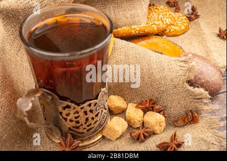 Ein facettiertes Glas Tee in einem Vintage-Cup-Halter, Sternanis und Stücke braunen Rohrzuckers auf einem Hintergrund aus strukturiertem Stoff. Nahaufnahme Stockfoto