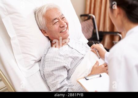 Asiatischer alter Mann, der im Bett liegt und mit junger Ärztin in der Krankenstation spricht Stockfoto