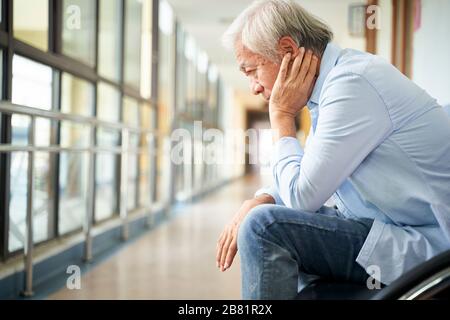 Trauriger und verwüsteter asiatischer Obermann, der allein in einem leeren Krankenhausflur sitzt, Kopf in der Hand Stockfoto