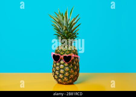 Ananas mit einer Paar rosafarbenen, herzförmigen Sonnenbrillen auf gelber Oberfläche vor blauem Hintergrund mit etwas Leerraum Stockfoto