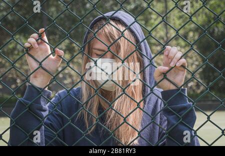 Geschlossener Rand. Quarantäne. Junge Frau, die eine Gesichtsmaske trägt, steht hinter dem Zaunrand. COVID-19 Coronavirus Infektion Pandemie-Virus. Stockfoto