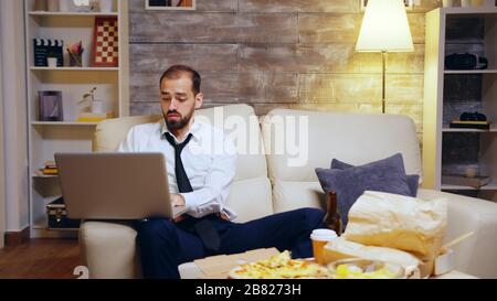 Der Geschäftsmann, der sein Bier aushat, während er von zu Hause aus mit dem Laptop arbeitete. Unternehmer im Anzug. Stockfoto