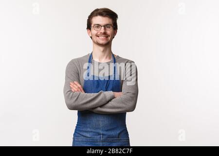 Taillenhoch Porträt des fröhlichen jungen männlichen Barista in der Schürze, überkreuzen Sie die Brust wie ein Profi, lächelnd glücklich, grüßt Kunden als stehend Stockfoto