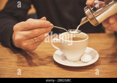 Junger Mann Hände halten, Gießen von Zucker aus Behälter in Kaffee. Selektive Fokus.man's Hand Gießen Zucker in Tasse mit schwarzem Kaffee.Süße heiße Getränk Stockfoto