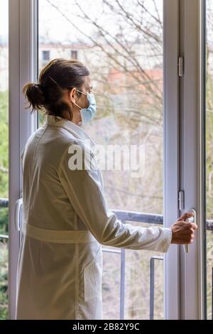 Eine Krankenschwester, die eine medizinische Gesichtsmaske und einen weißen Mantel trägt, steht untätig vor einem geschlossenen Fenster und starrt in den Weltraum. Stockfoto