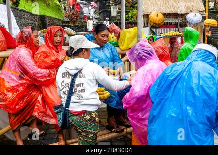 Lokale Frauen, die Sweetcorn/Corn an Besucher verkaufen, die Ponchos während EINER Regendusche tragen, die Batara Turun Kabeh Zeremonie, Besakih Tempel, Bali, Indonesien. Stockfoto