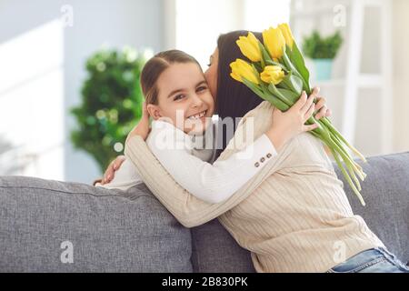 Glücklicher Muttertag. Tochter gratuliert Umarmungen, seine Mutter hält einen Blumenstrauß im Zimmer. Stockfoto