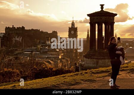 Edinburgh, Schottland, Großbritannien. März 2020. Farbenfroher Sonnenuntergang über dem Stadtzentrum. Mutter mit sehr jungem Baby, das in einer Tragseile geschnallt ist, nimmt ein selfie. Stockfoto