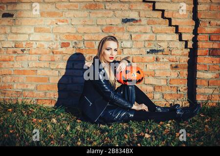 Moderne Halloween-Frau. Attraktive Hexe hält Kürbis "Trick oder Leckerei". Frau, die in Lederjacke und schwarzem Rock kleidet. Oktober halloween. Backsteine Stockfoto