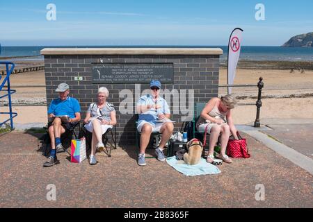 Vier Personen sitzen an der Strandpromenade von Llandudno, Wales, bei Sonnenschein. Stockfoto