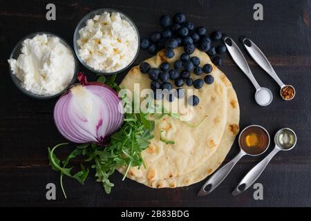 Zutaten für Blueberry, Feta und Honeykaramelisierte Zwiebel Naan Pizza: Naanische Fladenbrote, Arugula und andere Zutaten auf einem Holztisch Stockfoto