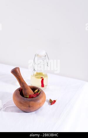 Holzmörtel, Pfirsich und rotes Chilipapier darin mit einer Flasche Olivenöl auf dem weißen Tischtuch gegen die hellgraue Wand. Stockfoto