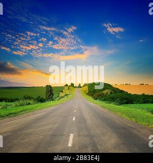 Sonnenaufgang über der malerischen Landschaft. Gerade Straße und die Felder leuchten Strahlen der Sonne. Stockfoto