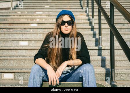 Urbane Frau mit Skate. Hipper-Mädchen mit Skateboard in der Stadt. Konzept für Extremsport und Emotionen. Alternativer Lebensstil. Modische Hüftmädchenhaltung Stockfoto