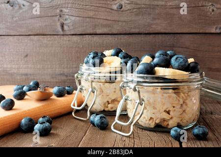 Oats über Nacht mit frischen Blaubeeren und Bananen in einem Schnappdeckelbecher auf rustikalem Holzhintergrund Stockfoto