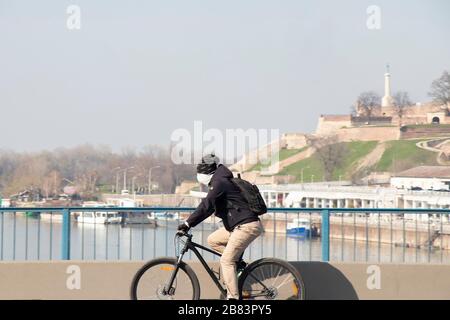 Belgrad, Serbien - 19. März 2020: Junge Männer, die Schutzmaske tragen, radeln über die Stadtbrücke mit der Festung Kalemegdan im Hintergrund, in der Zeit Stockfoto