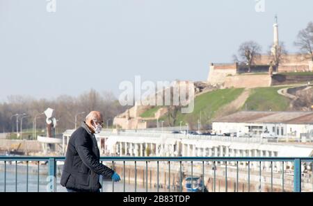 Belgrad, Serbien - 18. März 2020: Ältere Männer, die Schutzmasken tragen, die allein auf der Stadtbrücke mit der Festung Kalemegdan im Hintergrund in der ti spazieren gehen Stockfoto