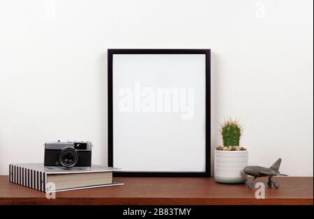 Verspotten Sie den schwarzen Rahmen mit Reisethema auf einem Regal oder Schreibtisch. Holzregal und weiße Wand. Ausrichtung des Hochformatrahmens. Stockfoto