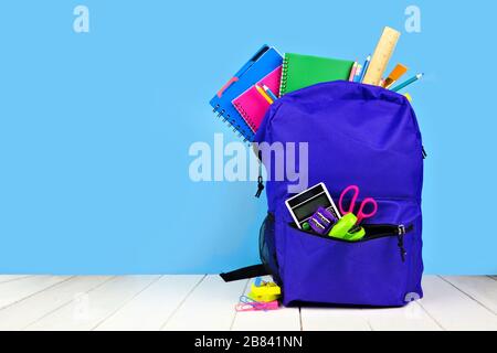 Violetter Rucksack voller Schulbedarf vor blauem Hintergrund. Zurück zum Schulkonzept. Kopierbereich. Stockfoto