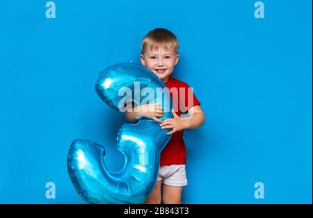 Kleiner süßer blonder Junge auf blauem Hintergrund mit Folienbeschichtung in baloonblauer Farbe. Alles gute zum Geburtstag drei Jahre alt Stockfoto