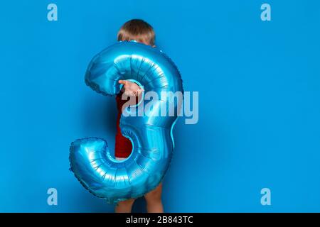 Kleiner süßer blonder Junge auf blauem Hintergrund mit Folienbeschichtung in baloonblauer Farbe. Alles gute zum Geburtstag drei Jahre alt Stockfoto