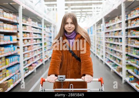 Junge Frau mit Lebensmittelkarre und Regalen mit Lebensmitteln in einem Laden Stockfoto