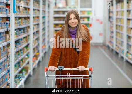 Junge Frau mit Lebensmittelkarre und Regalen mit Lebensmitteln in einem Laden Stockfoto