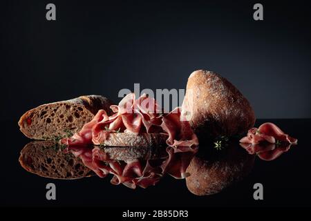 Prosciutto mit Ciabatta und Thymian auf schwarzem reflektierenden Hintergrund. Stockfoto