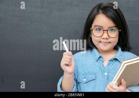Bildungskonzept. Ein Mädchen, das als Lehrerin eine Rolle spielt, Hand hält Kreide und Bücher. Sie trägt eine Brille, der Hintergrund ist eine Tafel. Es gibt ein B. Stockfoto