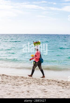 Frau aus der Region spaziert am weissgeschliffenen Strand von Isla Baru hinunter, um Obst aus einem Korb auf dem Kopf in der Nähe von Cartagena, Kolumbien zu verkaufen Stockfoto