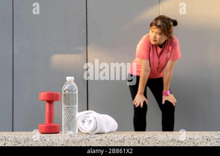 Fitnesskonzept. Frauen, die viel trainieren, stehen müde. Die Vorderseite hat eine Flasche reines Wasser, Hanteln, Handtücher. Stockfoto