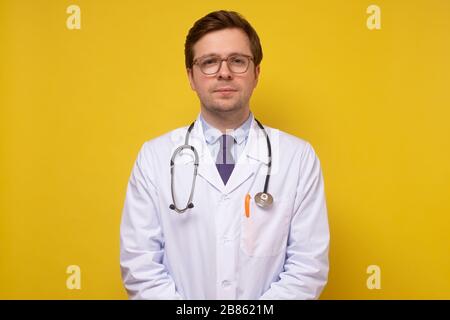 Ein Porträt eines medizinischen kaukasischen Arztes, der selbstsicher auf die Kamera lächelt, die vor gelbem Hintergrund posiert Stockfoto