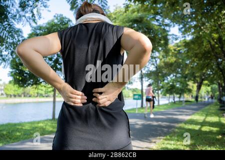 Konzept der Rückenschmerzen. Eine Frau, die mit einer Rückenverletzung in einem Sportanzug läuft, berührt den unteren Rückenmuskel, der auf dem Gehweg steht, der t erfasst Stockfoto