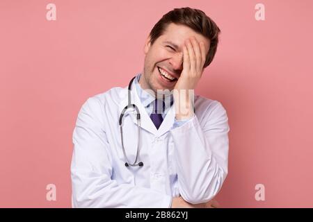 Junger kaukasischer Arzt, der in weißem Mantel aughing und Spaß hatte. Studio an pinkfarbener Wand Stockfoto