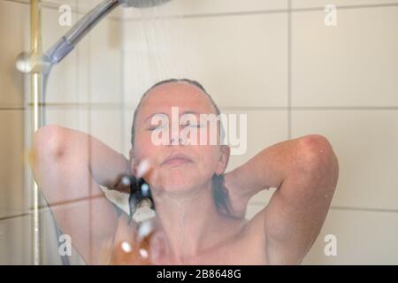 Frau, die eine dampfende heiße Dusche genießt, die ihr Haar unter fließendem Wasser mit geschlossenen Augen in einem Nahporträt durch die Glaskabine in abspült Stockfoto