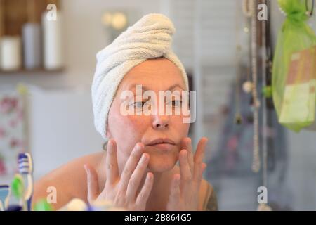 Frau, die ein Handtuch auf ihrem nassen Haar trägt, nachdem sie in einem Badezimmerspiegel in einem persönlichen Hygienekonzept ihr Gesicht unter die Haut geduschen hat Stockfoto