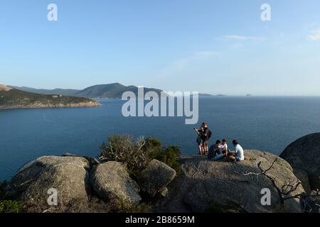 Eine Gruppe junger Männer spielt Karten auf einem Hügel mit Blick auf die Bucht, während ein junges Paar Fotos von der Aussicht nimmt Stockfoto