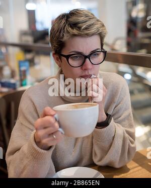 Frau, die einen Pullover aus Elfenbein und eine schwarze Brille trägt, ein Telefon spießt, ihre Nase wischt und nach dem Einkaufen in einem Café eine Tasse Tee hat Stockfoto