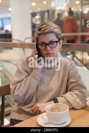 Frau, die einen Pullover aus Elfenbein und eine schwarze Brille trägt, ein Telefon spießt, ihre Nase wischt und nach dem Einkaufen in einem Café eine Tasse Tee hat Stockfoto