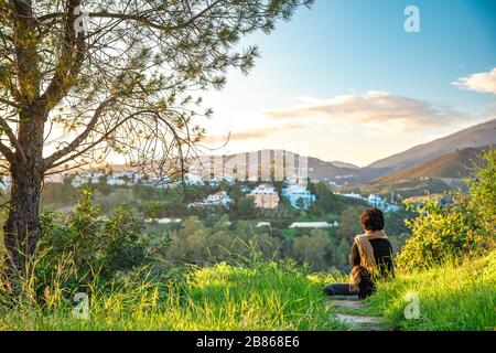 Frau, die sich bei Sonnenuntergang auf einem grünen Hügel erholt und meditiert, mit wunderschönem Blick auf die Landschaft und die Berge Stockfoto