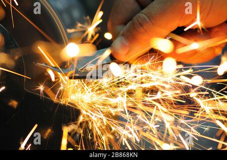 Der Mann hält Details in der Hand und arbeitet mit Geräten zum Schneiden von Metall, helle feurige Funken fliegen auseinander, Nahaufnahme Foto Stockfoto